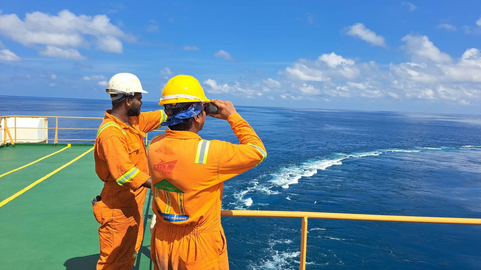 Two workers in orange coveralls and safety gear looking out to sea from a ship's deck.