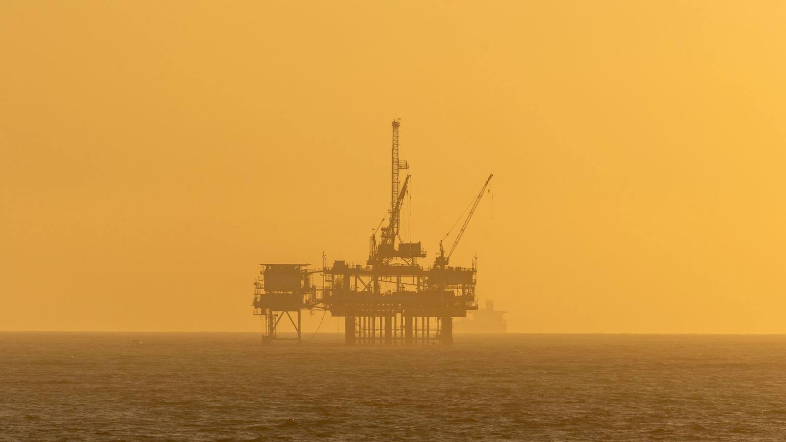 An offshore oil rig silhouetted against a golden sunset over the ocean.