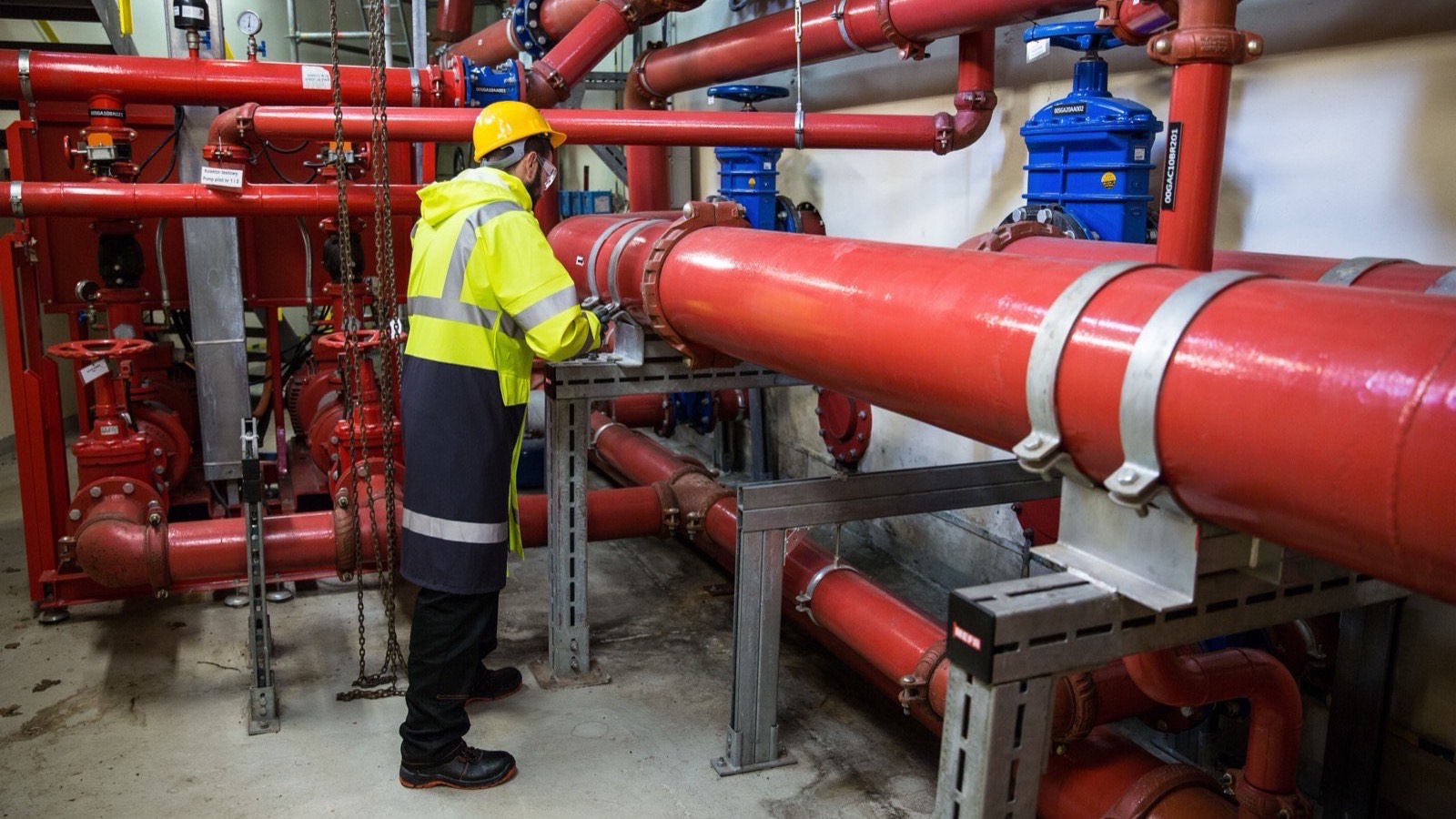 A worker in a hard hat and high-visibility coat inspecting pipework and valves