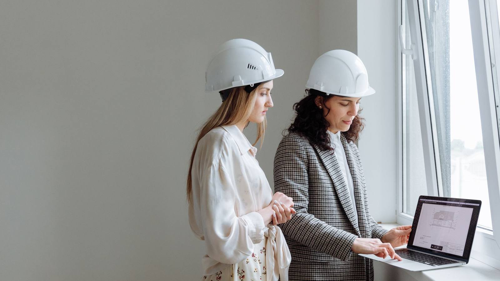 Two women in hard hats reviewing plans on a laptop by a window.