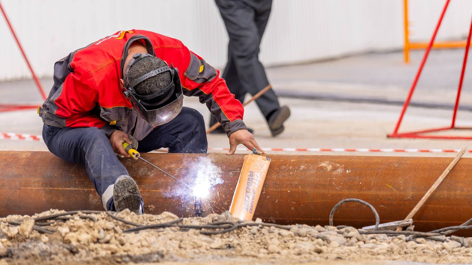 A worker in safety gear welding a steel pipe at an outdoor construction site.